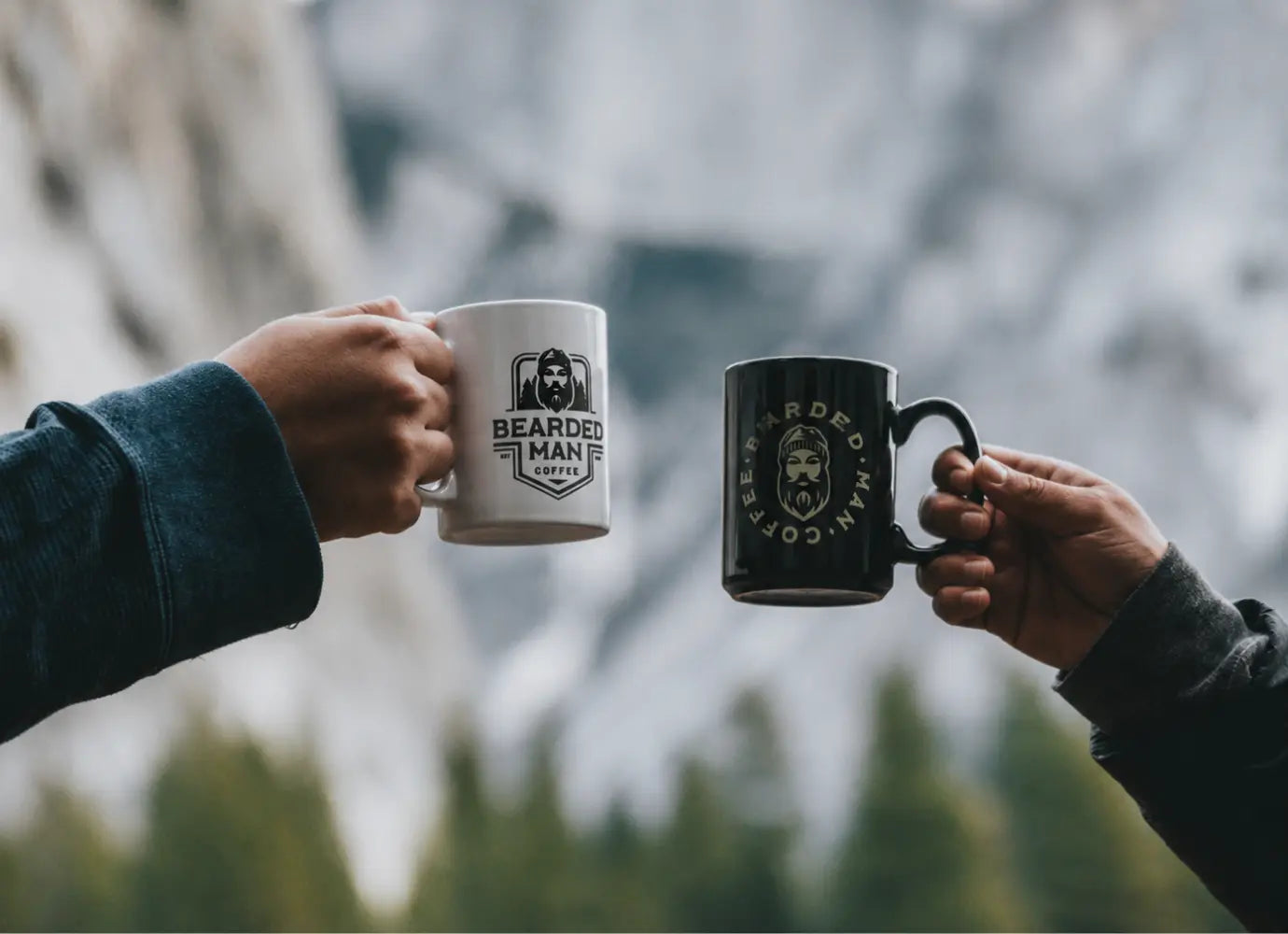 Two people holding Bearded Man Coffee mugs together in front of a blurred mountain landscape.